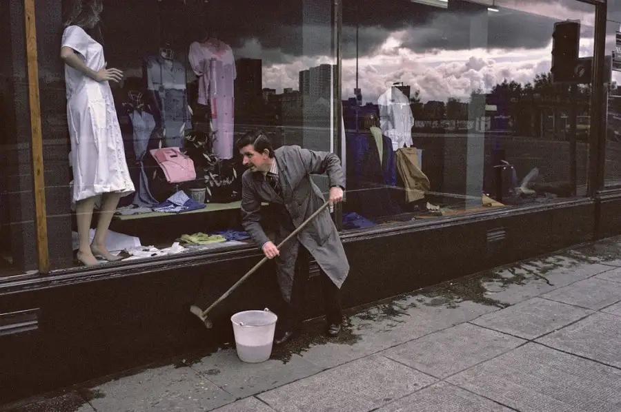 A man in a grey trench coat is mopping the sidewalk outside a clothing store, with a bucket beside him. The store window…