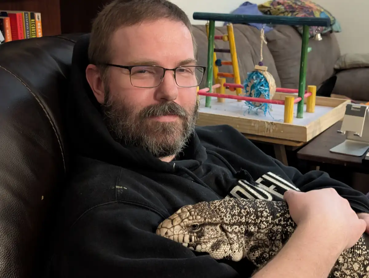 A man with glasses and a beard sits in a black leather chair, holding a large, patterned lizard (likely a bearded dragon or…