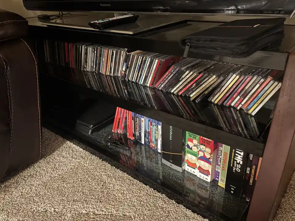 A glass TV stand holds a large collection of DVDs and CDs, with the top shelf filled with CDs standing upright and the…