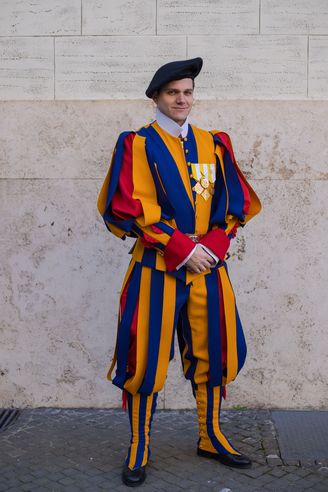 A man in a colorful, vertically striped blue and yellow ceremonial uniform with a black beret stands against a stone wall…