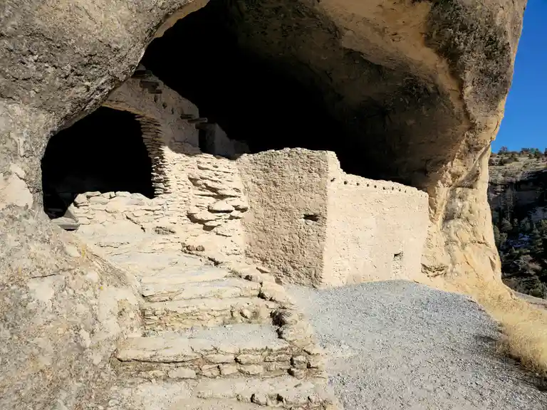The image shows ancient stone ruins built into the side of a large natural rock overhang, with stone steps leading up to…