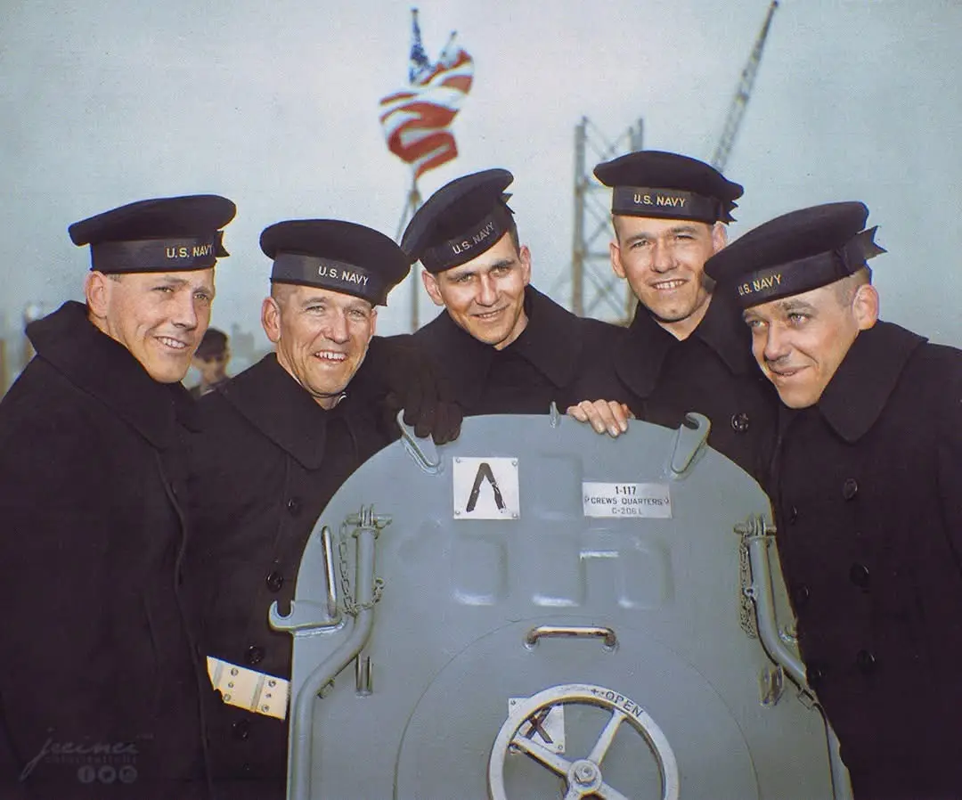Five smiling U.S. Navy sailors in dark uniforms and caps pose around a large gray hatch labeled 'CREWS QUARTERS' with a…