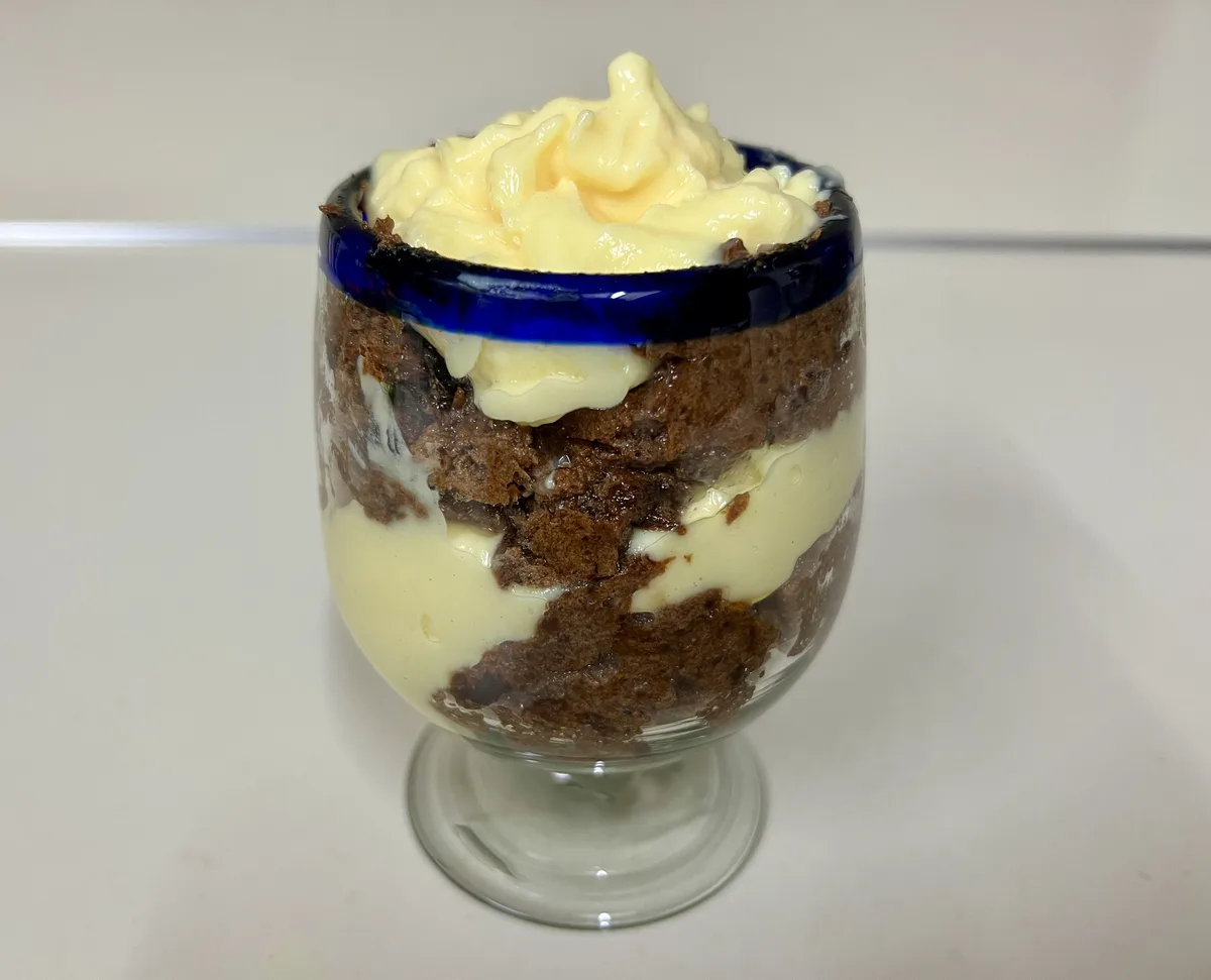 A layered dessert in a clear glass cup with a blue rim, featuring alternating layers of brown cake and creamy yellow custard…