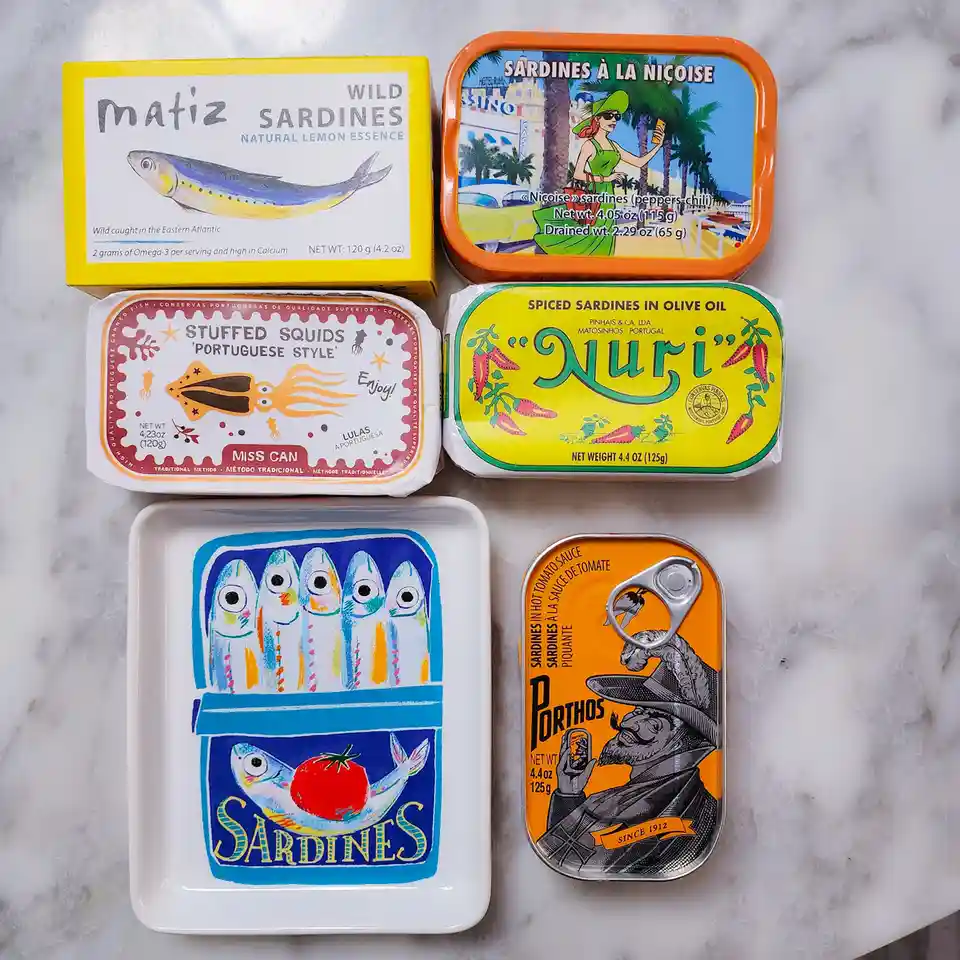 Six colorful canned fish and seafood products are arranged on a marble surface, including sardines, stuffed squids, and a…