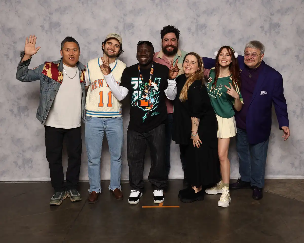Seven people pose together in front of a gray backdrop, smiling and making peace signs. The person in the center wears a…