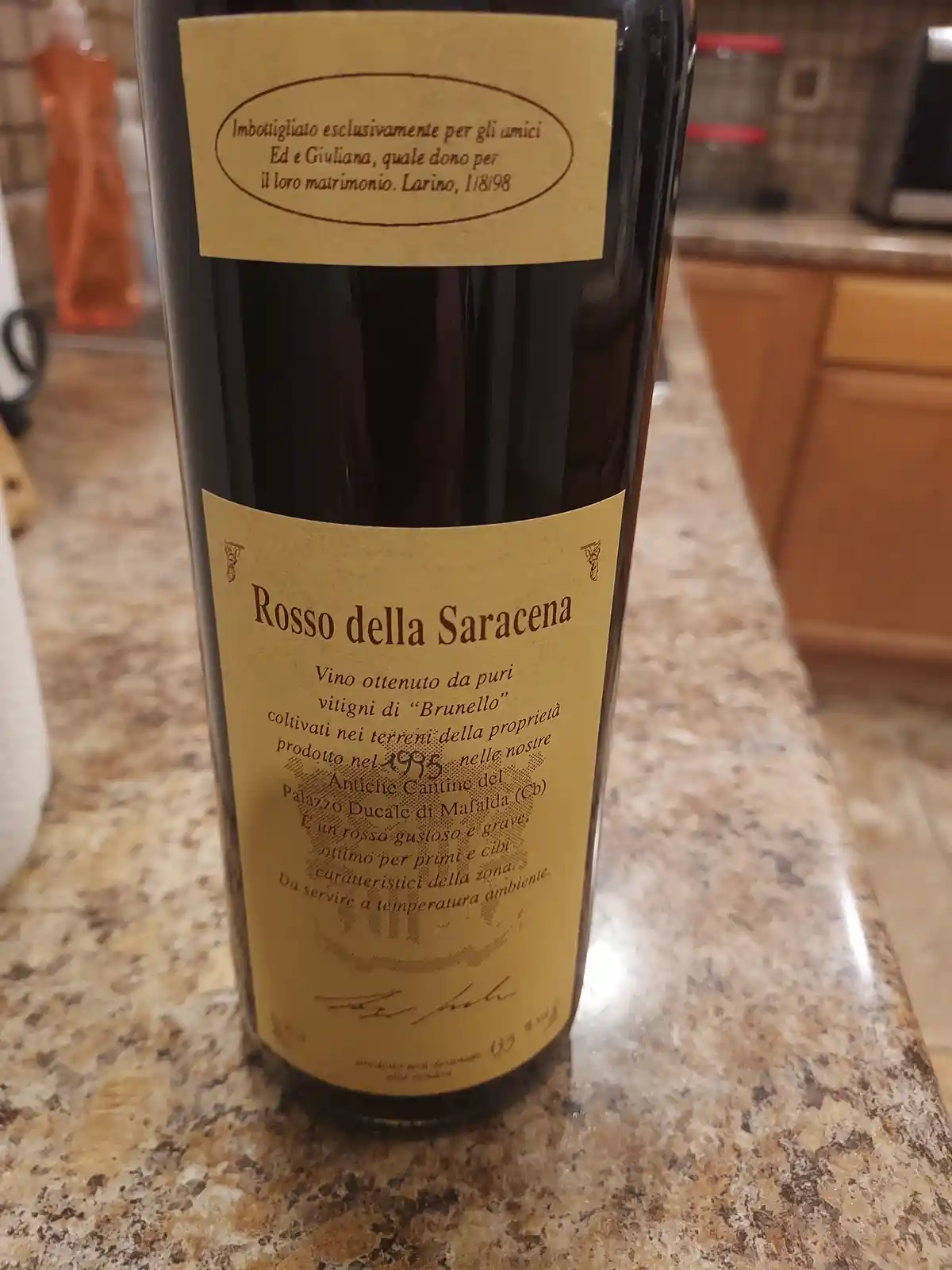 A close-up photo of a wine bottle labeled 'Rosso della Saracena' sitting on a granite countertop. The bottle has a gold…