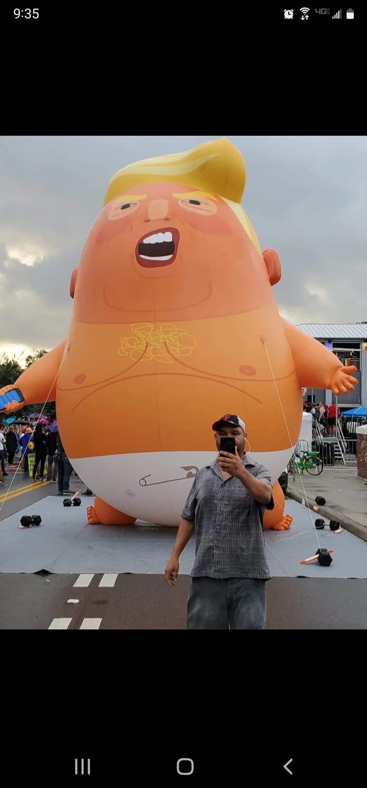 A man takes a selfie in front of a giant inflatable baby balloon depicting a cartoonish figure with a large head, orange…