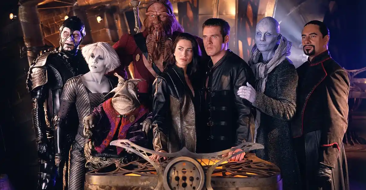A promotional group photo of the cast of Farscape, featuring diverse alien and human characters in sci-fi costumes, standing…