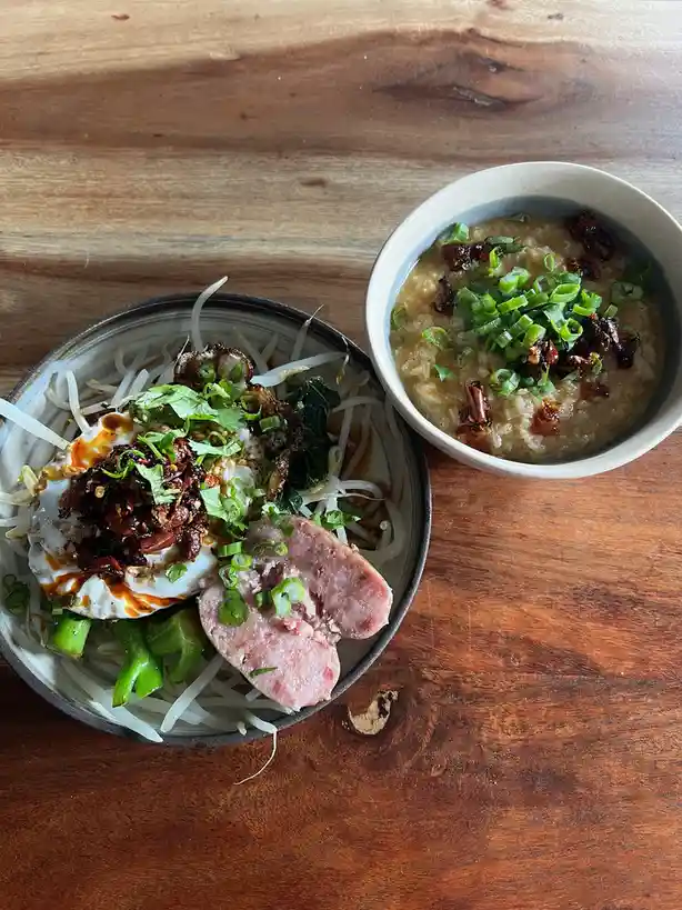 Two bowls of food sit on a wooden table: one contains noodles topped with a fried egg, bean sprouts, herbs, and chili oil…