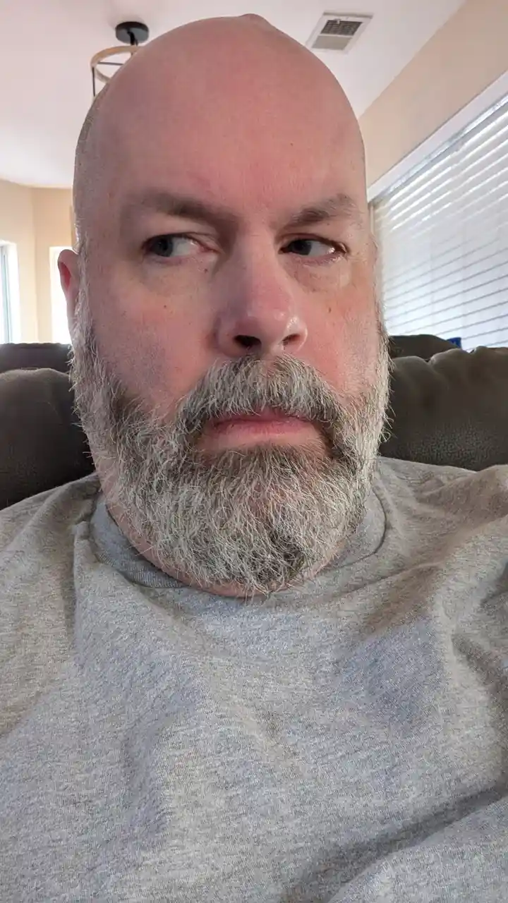 A bald man with a gray beard and mustache sits on a couch, looking sideways with a deadpan expression. He wears a gray…