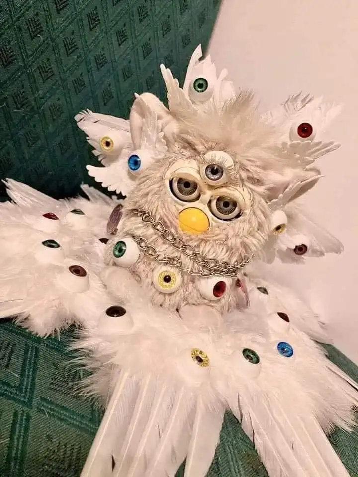 A heavily modified, white, fluffy Furby toy is shown sitting on a green patterned couch. Its body and wings are covered in…