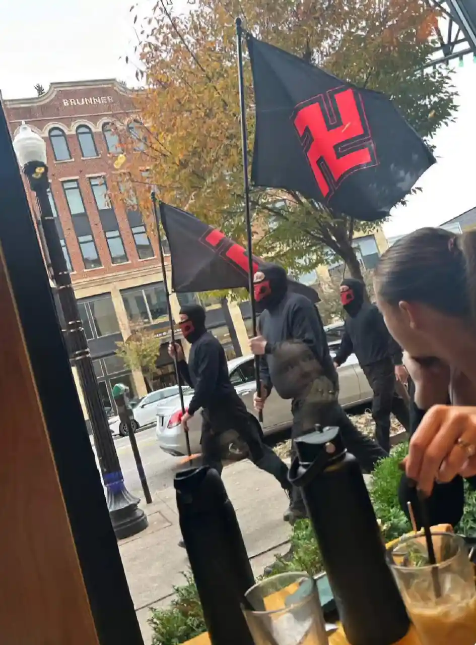 A person is seen from behind, sitting at a cafe table with drinks and water bottles, looking out the window at a group of…