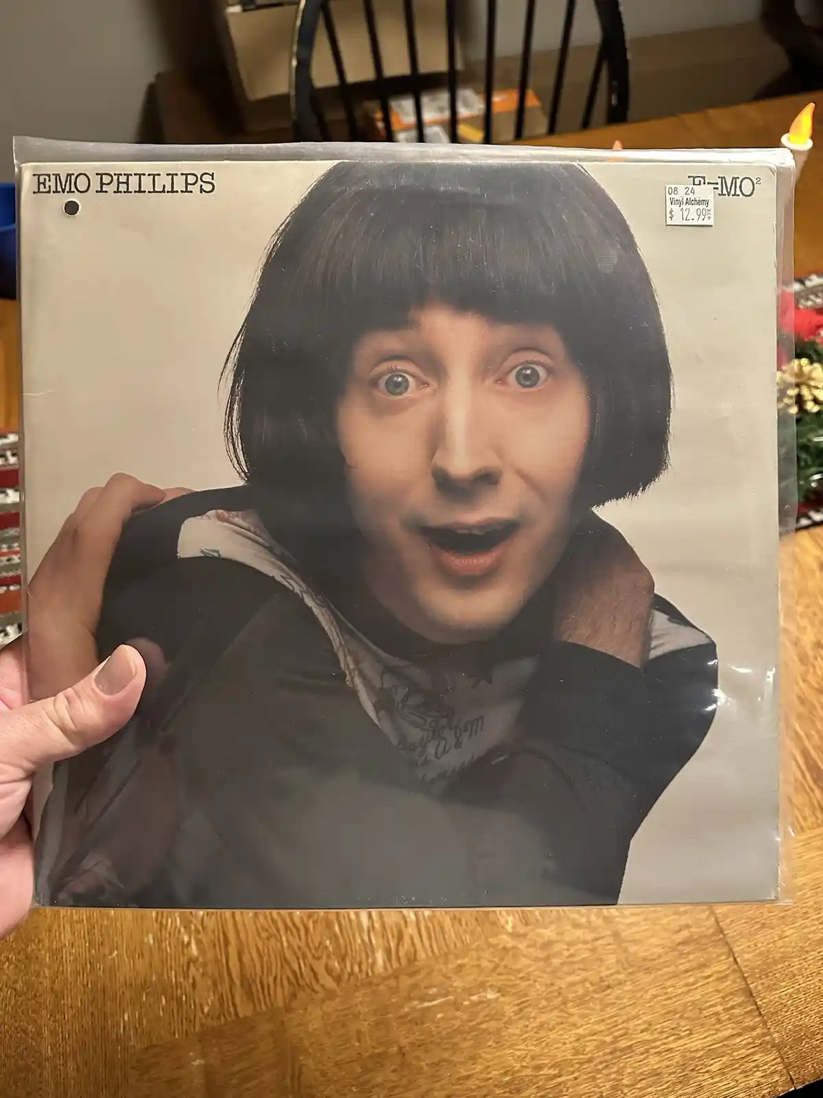 A person holds up a vinyl record album cover featuring comedian Emo Philips with a surprised expression, wearing a black bob…