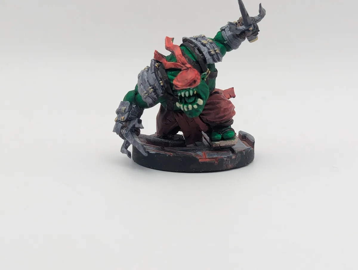 A painted miniature of a green-skinned, muscular orc-like warrior with red bandana and armor, wielding a spiked weapon and…