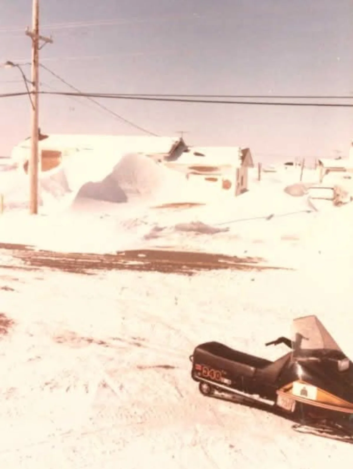 A vintage, faded photograph shows a snowmobile parked in a snowy, rural landscape with houses and a utility pole in the…