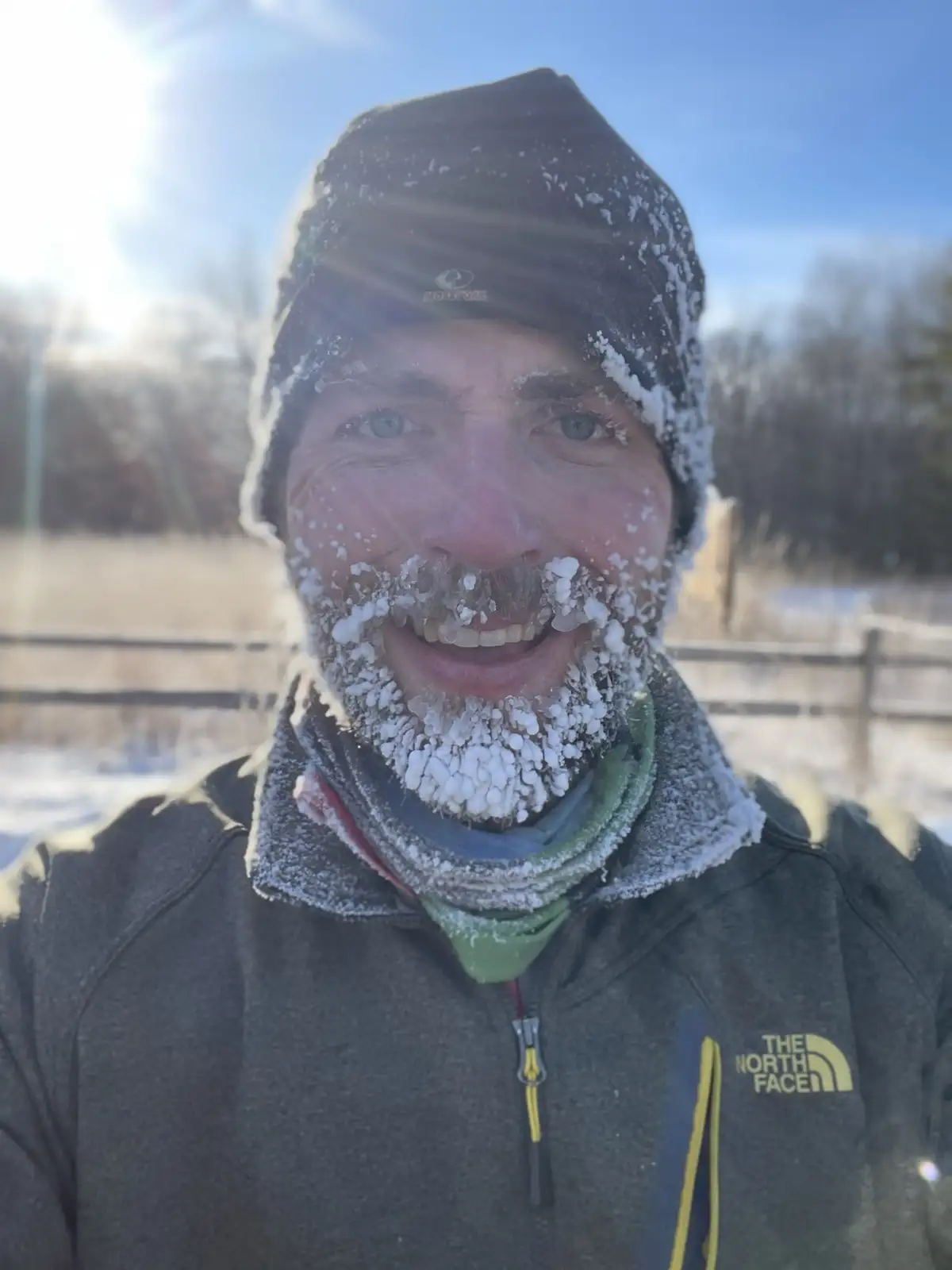 A man with a frost-covered beard and hat smiles at the camera outdoors in winter, wearing a North Face jacket and scarf…