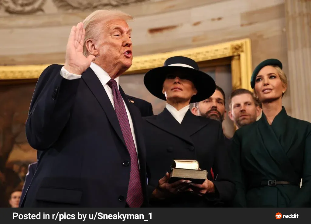 A man in a dark suit and tie raises his right hand while speaking, standing beside a woman in a black coat and hat holding a…