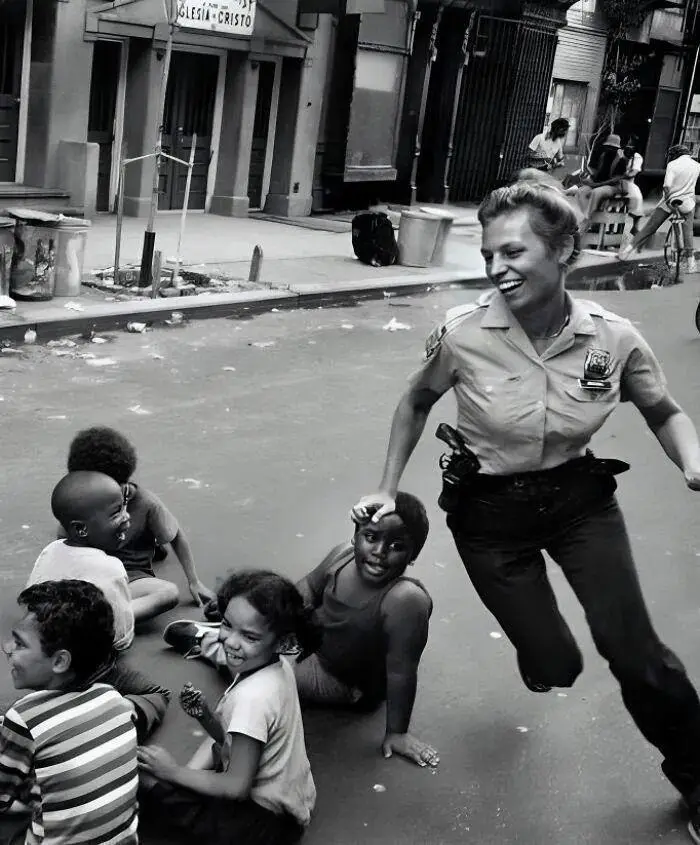 A black-and-white photograph shows a smiling police officer running toward a group of children sitting on a street. She has…