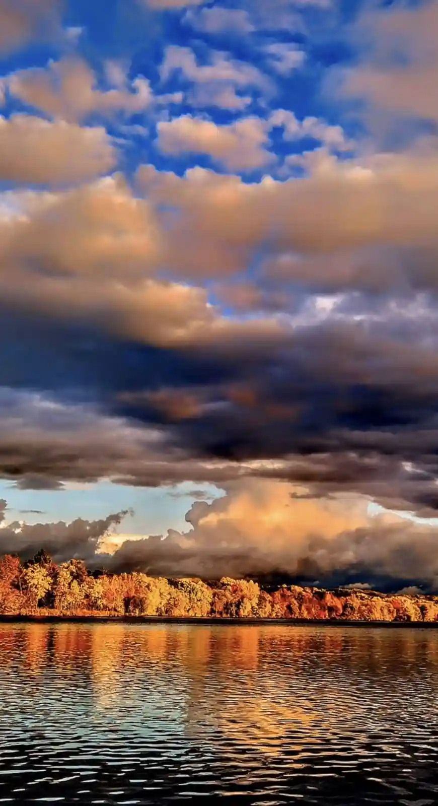 A dramatic landscape photo shows a lake reflecting a sky filled with golden and dark clouds at sunset or sunrise, with a…