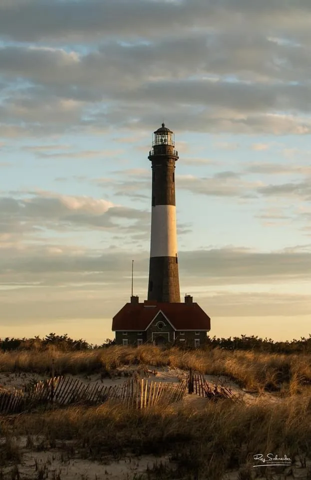 A tall, black-and-white striped lighthouse stands on a sandy dune with a red-roofed building at its base, under a cloudy sky…