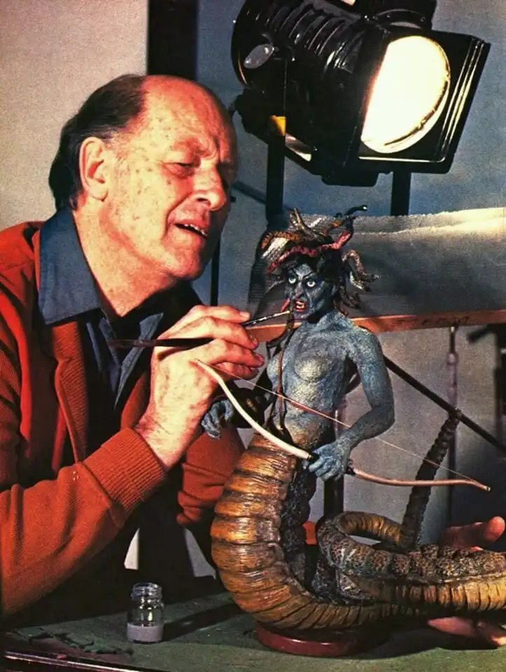 A man in a red sweater is painting a detailed blue-skinned monster sculpture with a bow and arrow, under a bright studio…