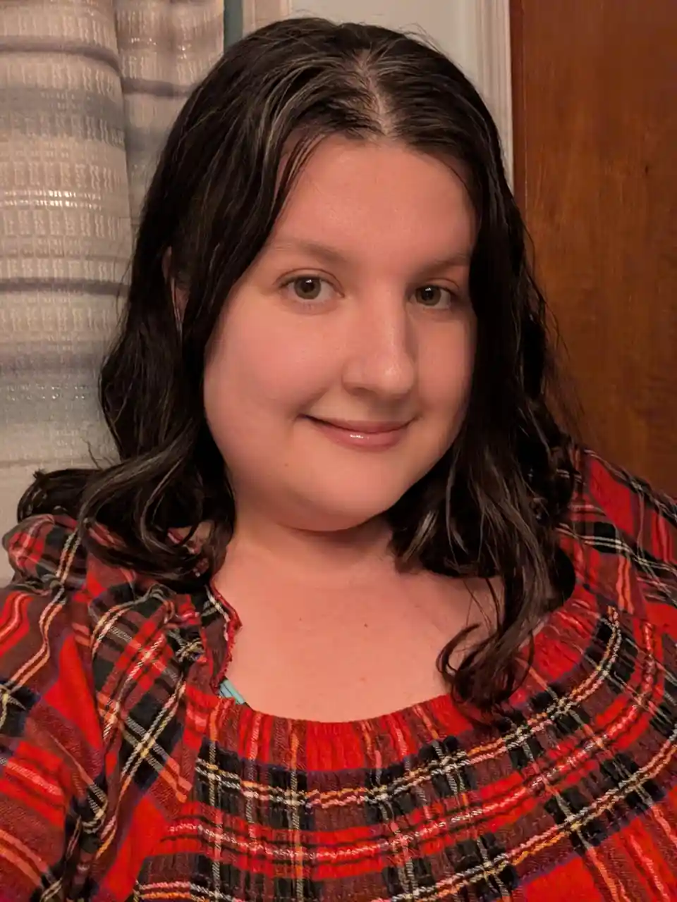 A person with dark, wavy hair and a subtle smile wears a red, black, and orange plaid shirt. Behind them is a striped…