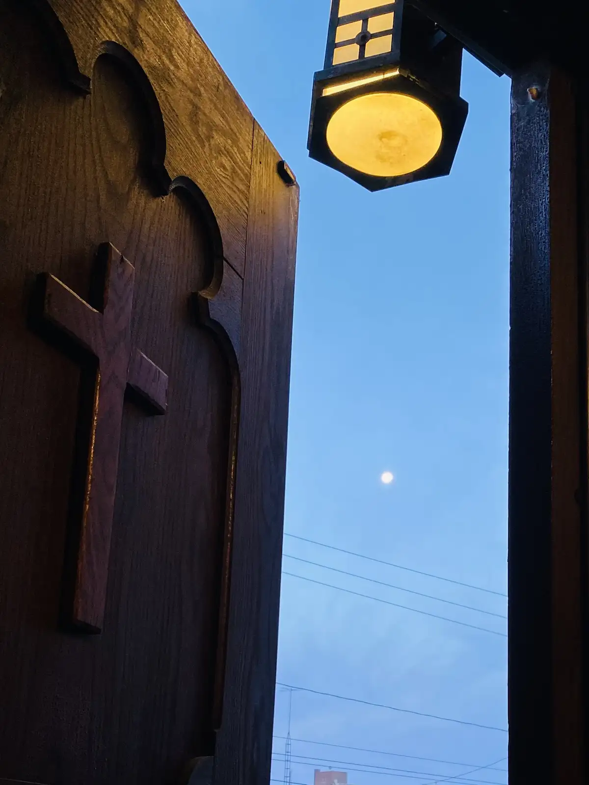 A low-angle photo shows a wooden church door with a cross, next to a lit streetlamp, framing a pale moon in a twilight sky…