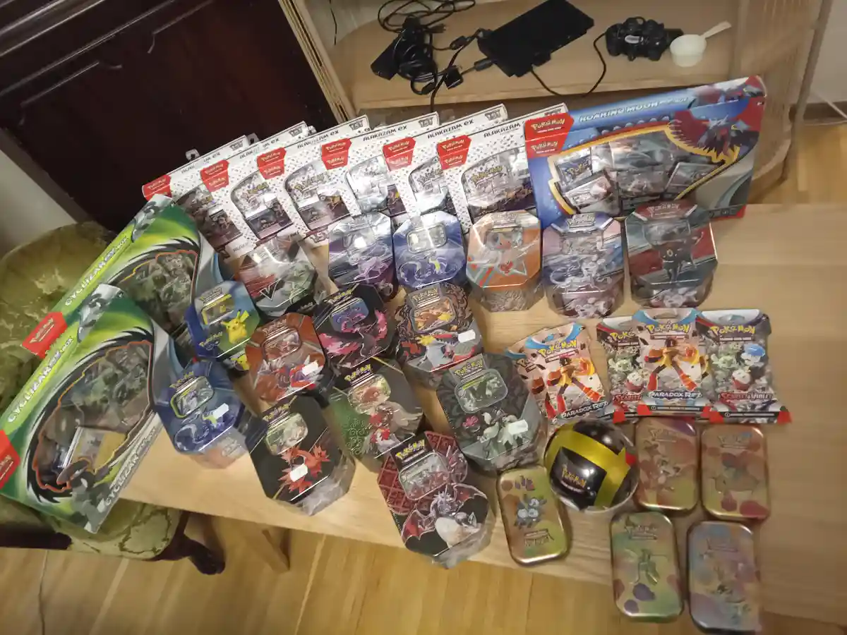 A wooden table is covered with a large collection of Pokémon trading card game products, including multiple 'Alakazam EX'…