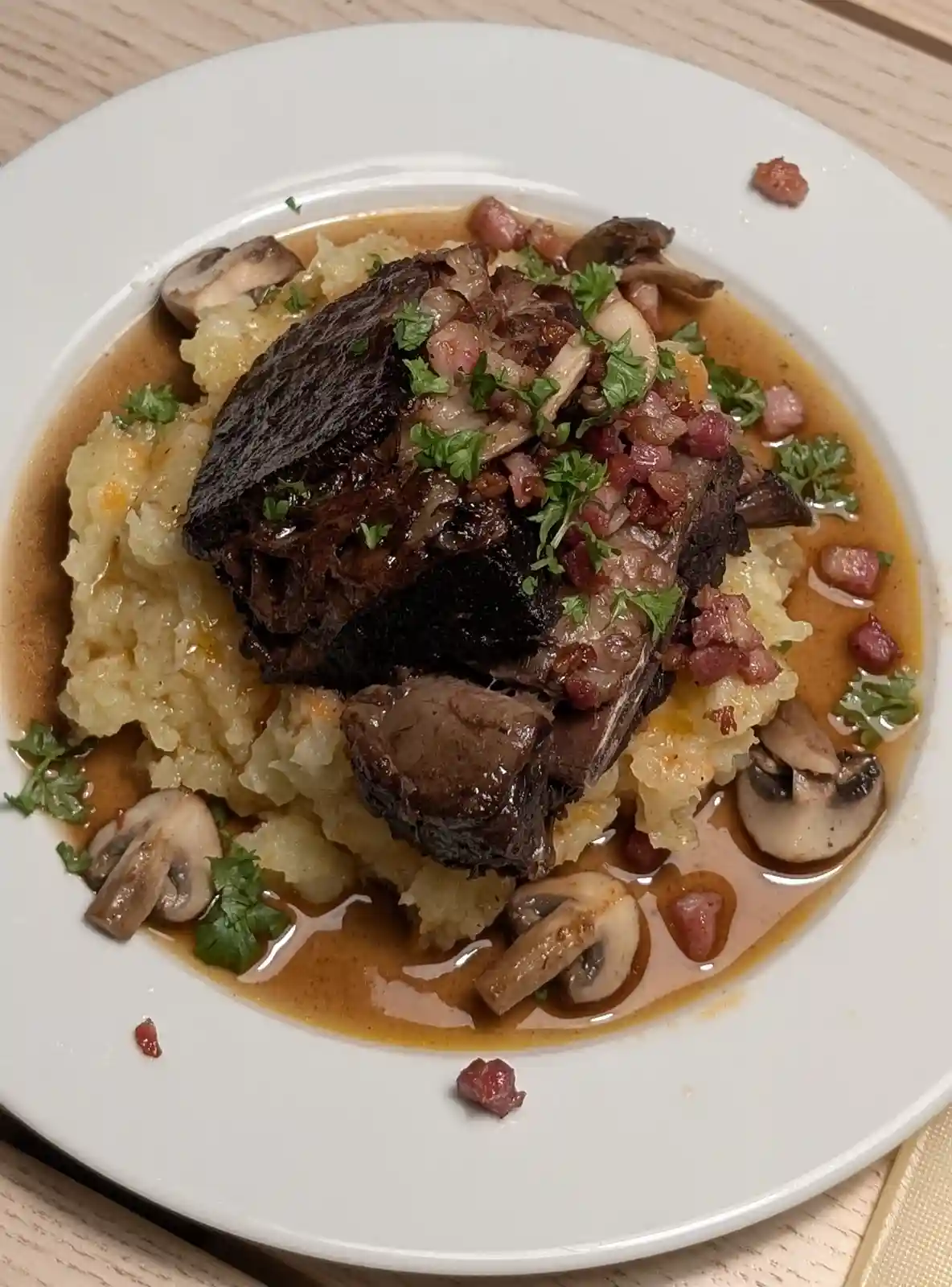 A close-up photo of a plated gourmet dish featuring a large, dark seared meat roast atop a bed of mashed potatoes, garnished…