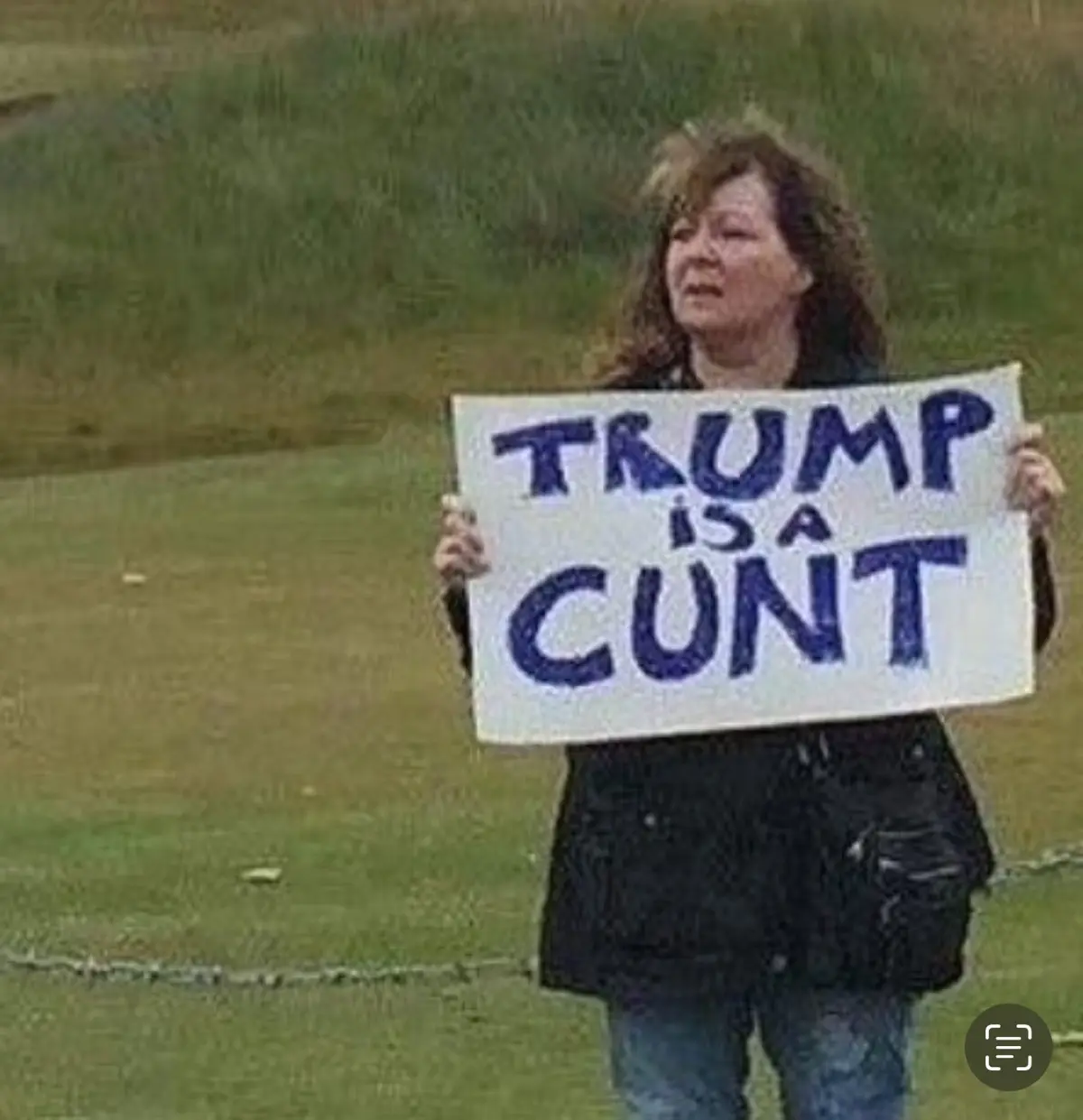 A woman with curly brown hair stands outdoors in a grassy field, holding a handmade white sign with bold blue handwritten…