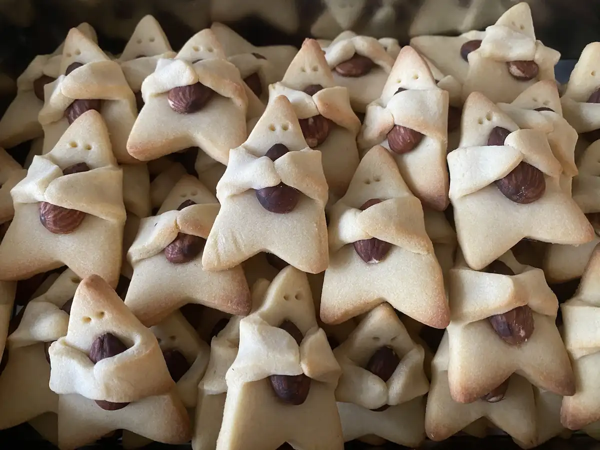 A close-up shot shows dozens of star-shaped, golden-brown cookies arranged in a pile. Each cookie has a whole almond placed…
