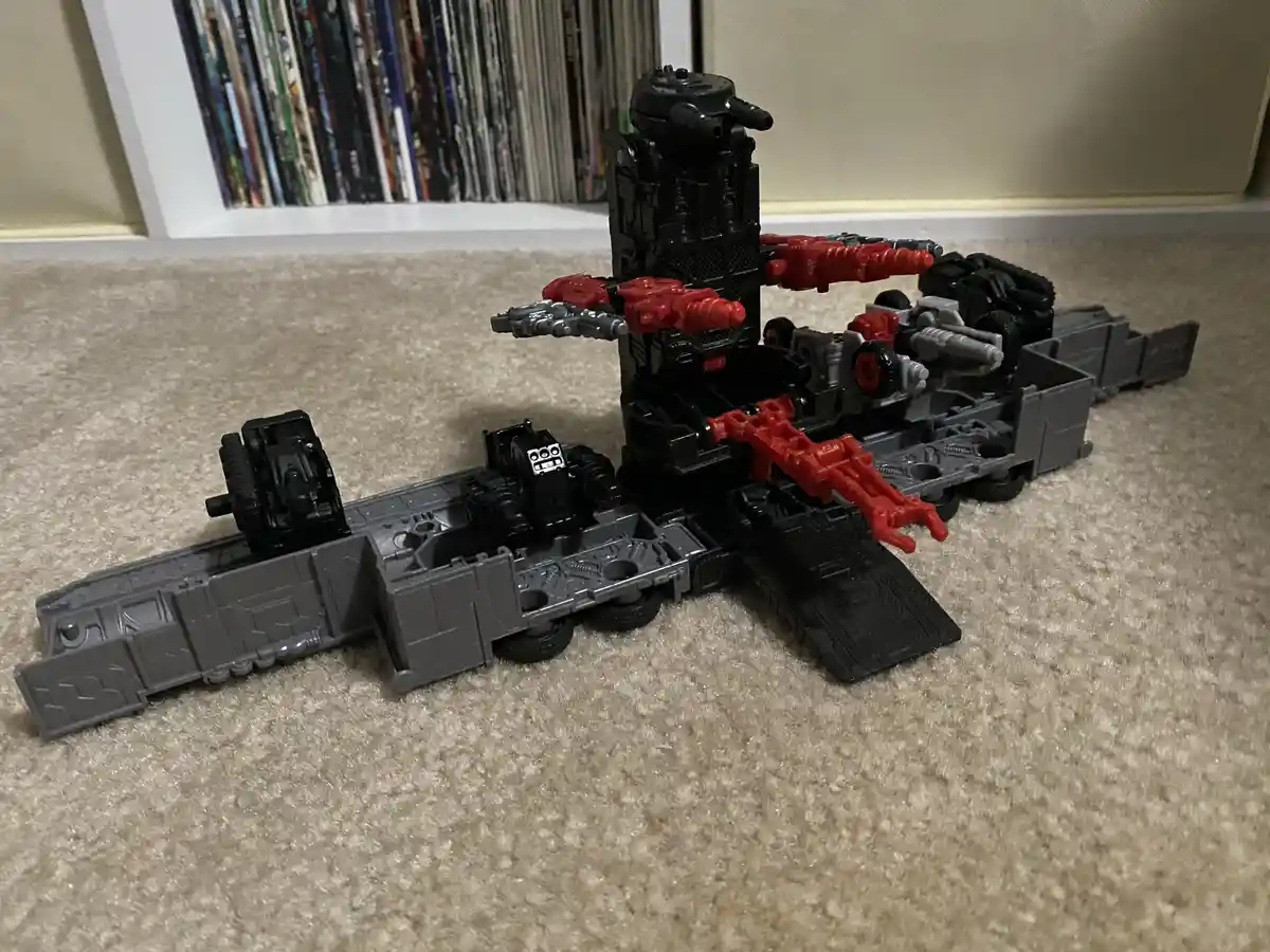A large, complex toy robot, primarily black and red with grey mechanical parts, is shown in a partially transformed state on…