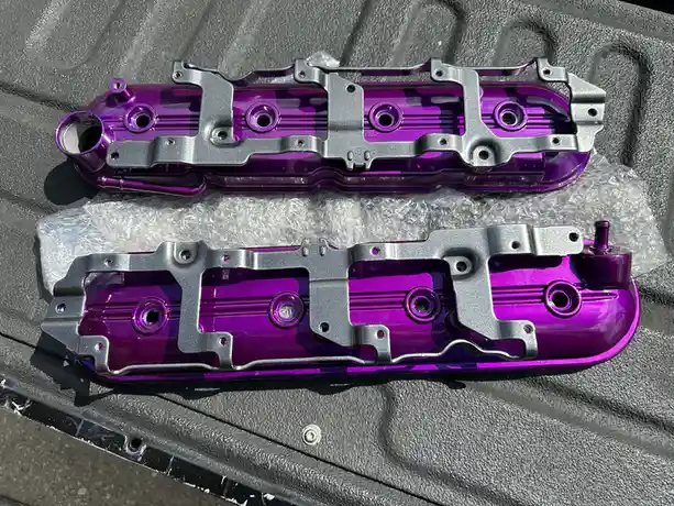 Two shiny, custom-painted purple and silver automotive valve covers rest on bubble wrap inside a textured gray case. The…