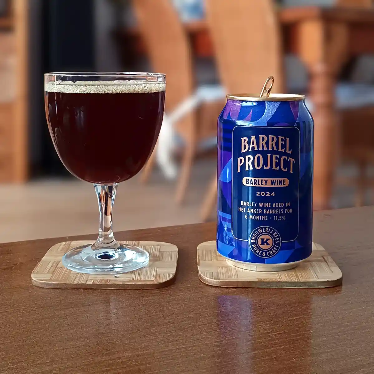 A blue can of 'Barrel Project Barley Wine 2024' by Brouwerij Kees sits next to a glass of dark, foamy beer on wooden…
