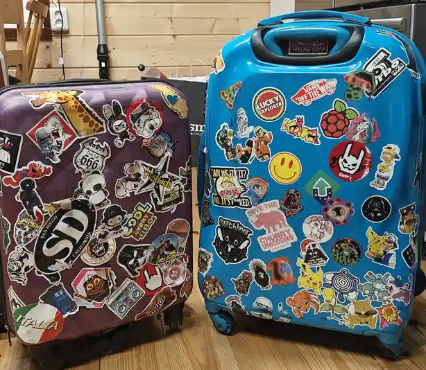 Two rolling suitcases, one purple and one blue, are covered in dozens of colorful stickers featuring pop culture icons…