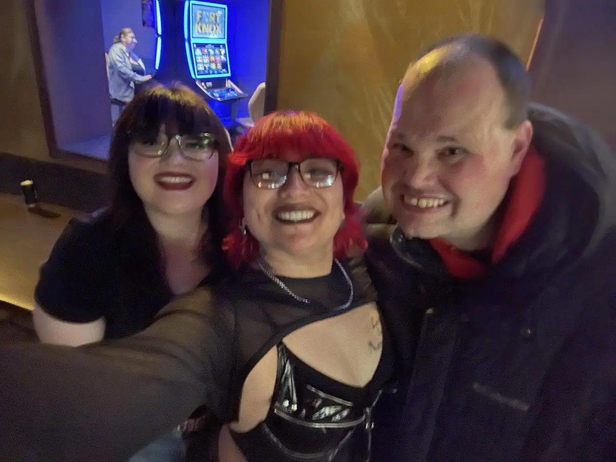 Three people are taking a selfie in a casino, with a slot machine labeled 'FORT KNOX' visible in the background. The person…