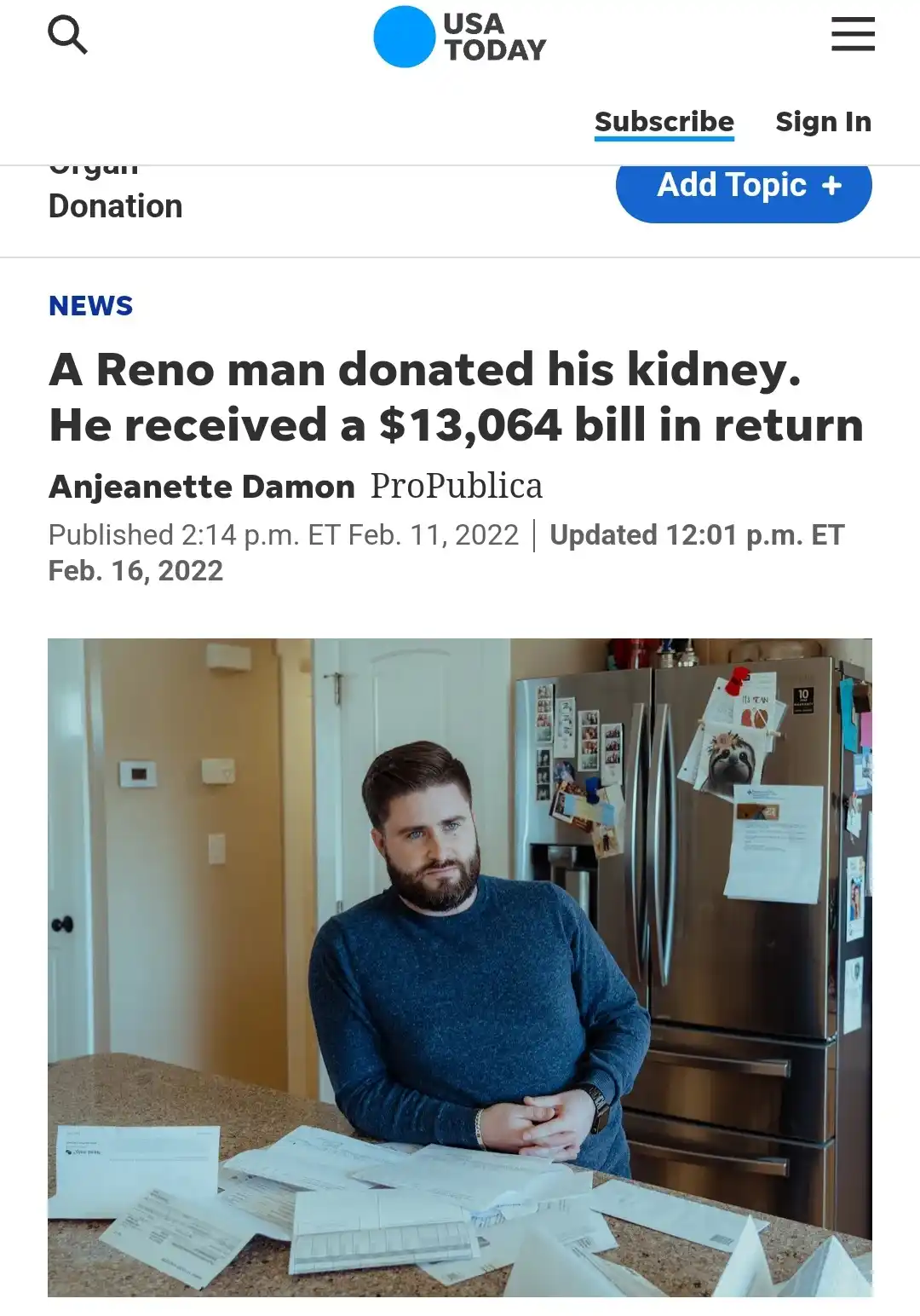 A man with a beard and blue sweater sits at a kitchen counter surrounded by scattered bills and envelopes, looking weary…