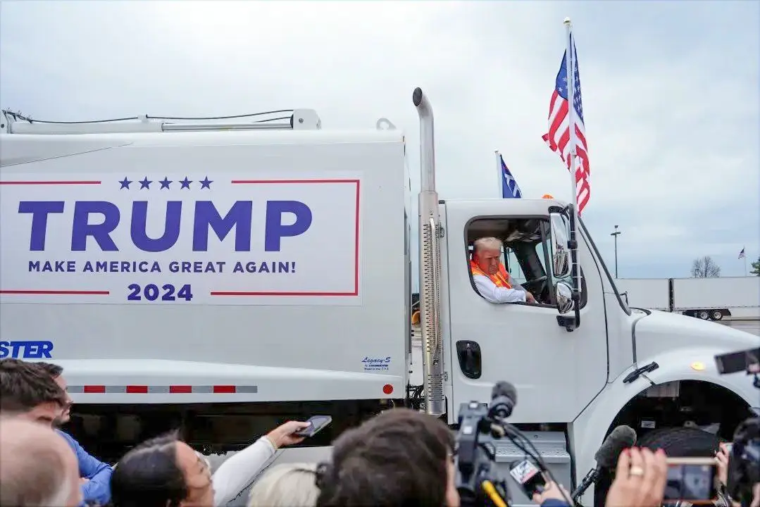 A man wearing an orange safety vest sits in the driver's seat of a white semi-truck, which has a large political campaign…