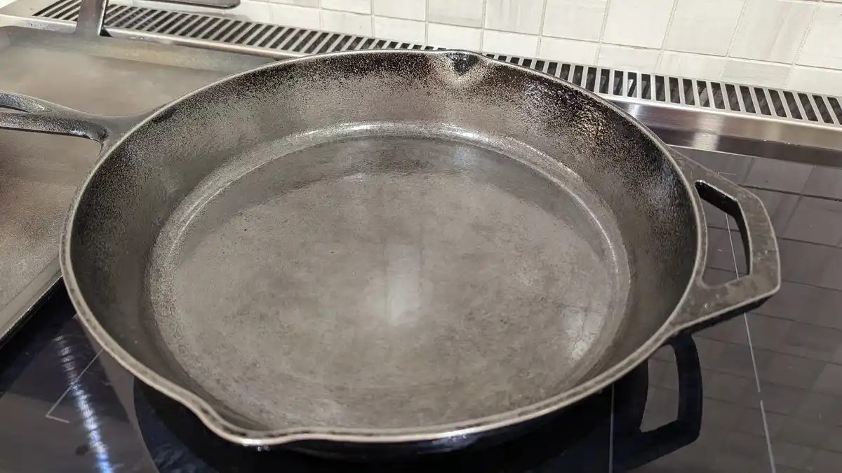 A close-up photo shows a well-seasoned, empty cast iron skillet resting on a stovetop. The skillet has a dark, slightly worn…