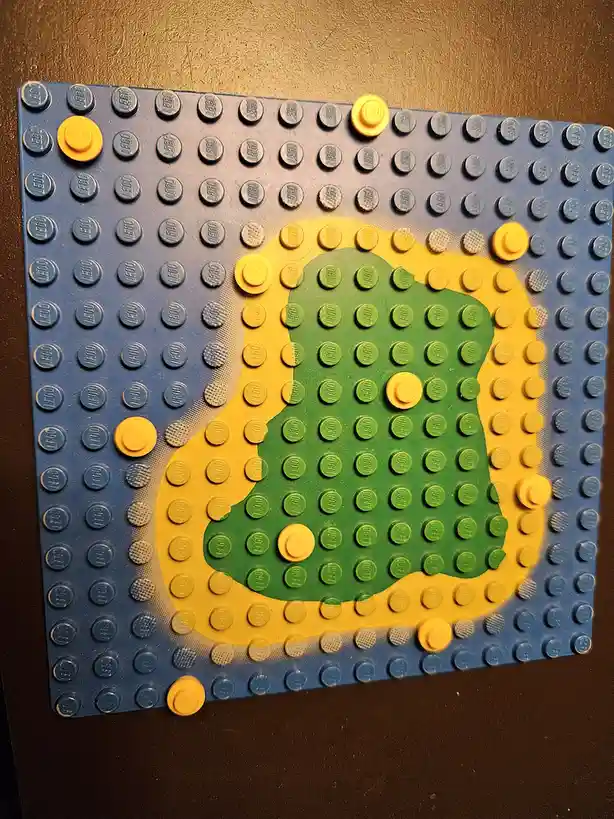 A blue Lego baseplate has a green and yellow shape resembling a stylized face or mask drawn with spray paint, with a few…