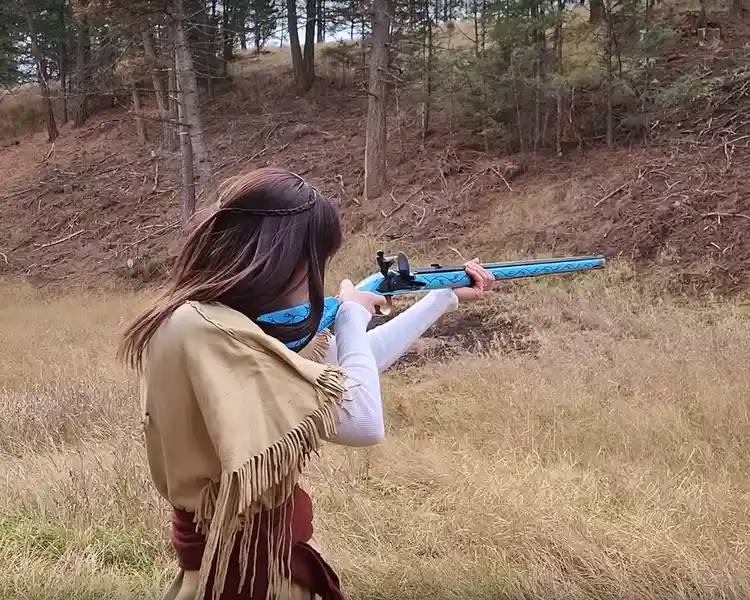 A person with long brown hair, wearing a tan fringed poncho over a white shirt and a blue patterned bandana, is aiming a…
