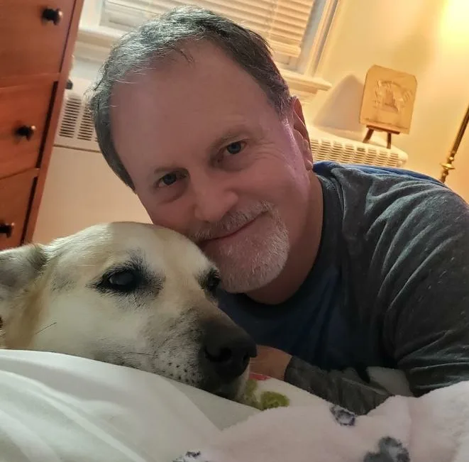 A man with gray hair and a goatee smiles warmly while lying in bed next to a light-colored dog whose head rests on a…