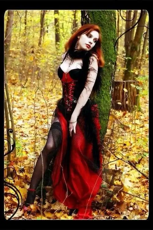 A woman with red hair and pale makeup stands leaning against a mossy tree in an autumn forest, surrounded by fallen yellow…