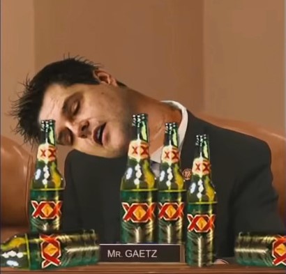 A man in a suit is slumped over a desk, appearing intoxicated, with multiple bottles of XX beer arranged in front of him. A…
