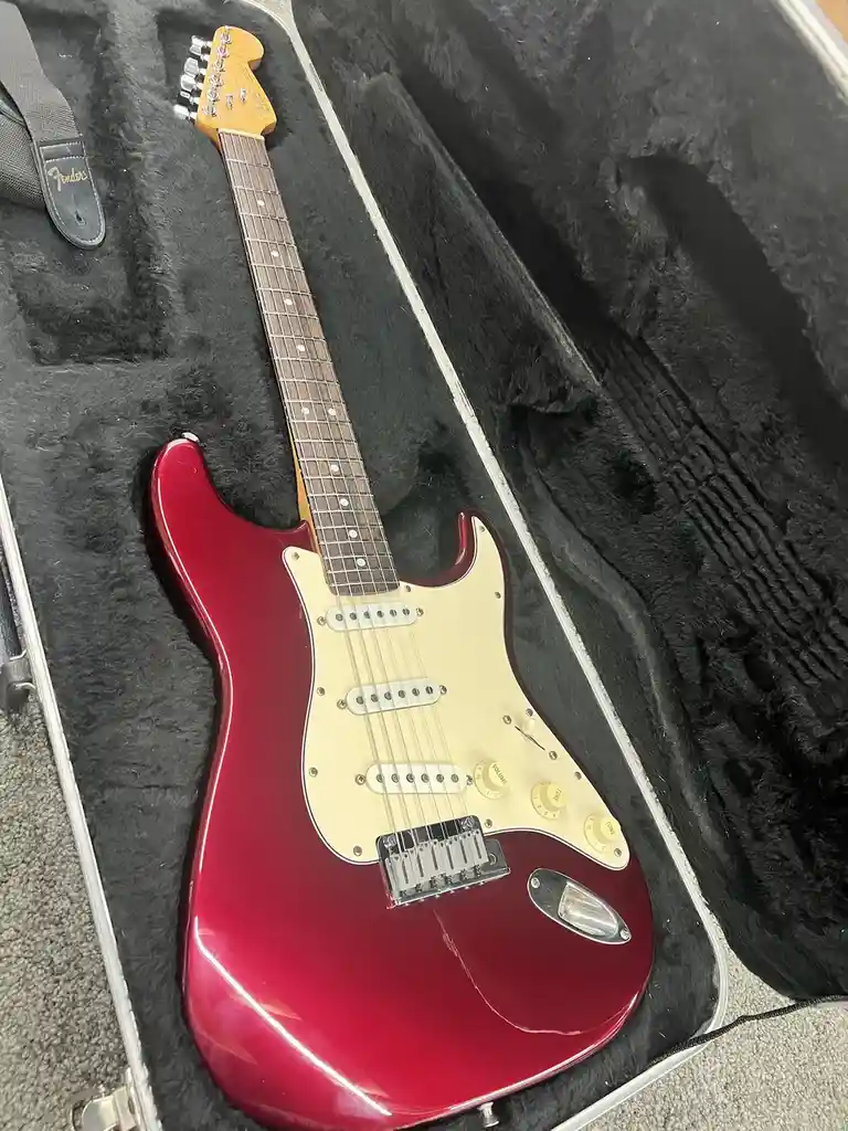 A glossy red Fender Stratocaster electric guitar rests inside its open black-lined hard case, with a Fender strap visible…