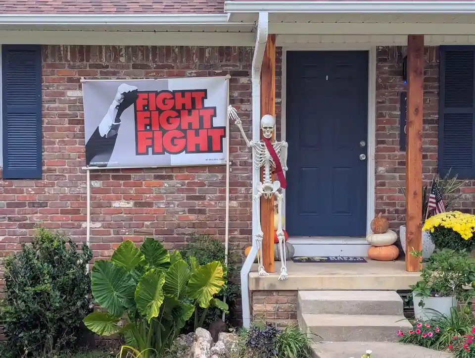A Halloween-decorated brick house porch features a skeleton wearing a red tie, standing beside a blue door. A large banner…