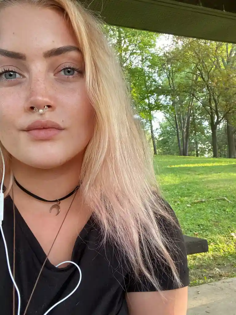A young woman with blonde hair, freckles, and a nose ring looks directly at the camera with a neutral expression while…