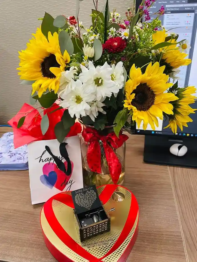 A desk setup features a large, vibrant bouquet of sunflowers and white daisies in a glass vase tied with a red bow, next to…