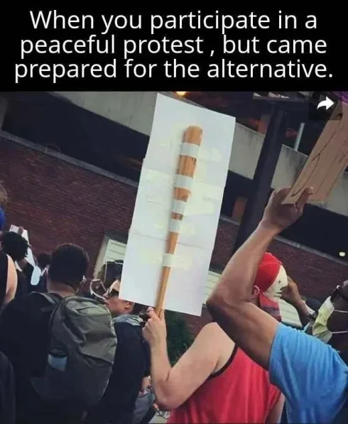 A person at a protest holds up a large white sign taped to a wooden baseball bat, while other protesters are visible in the…