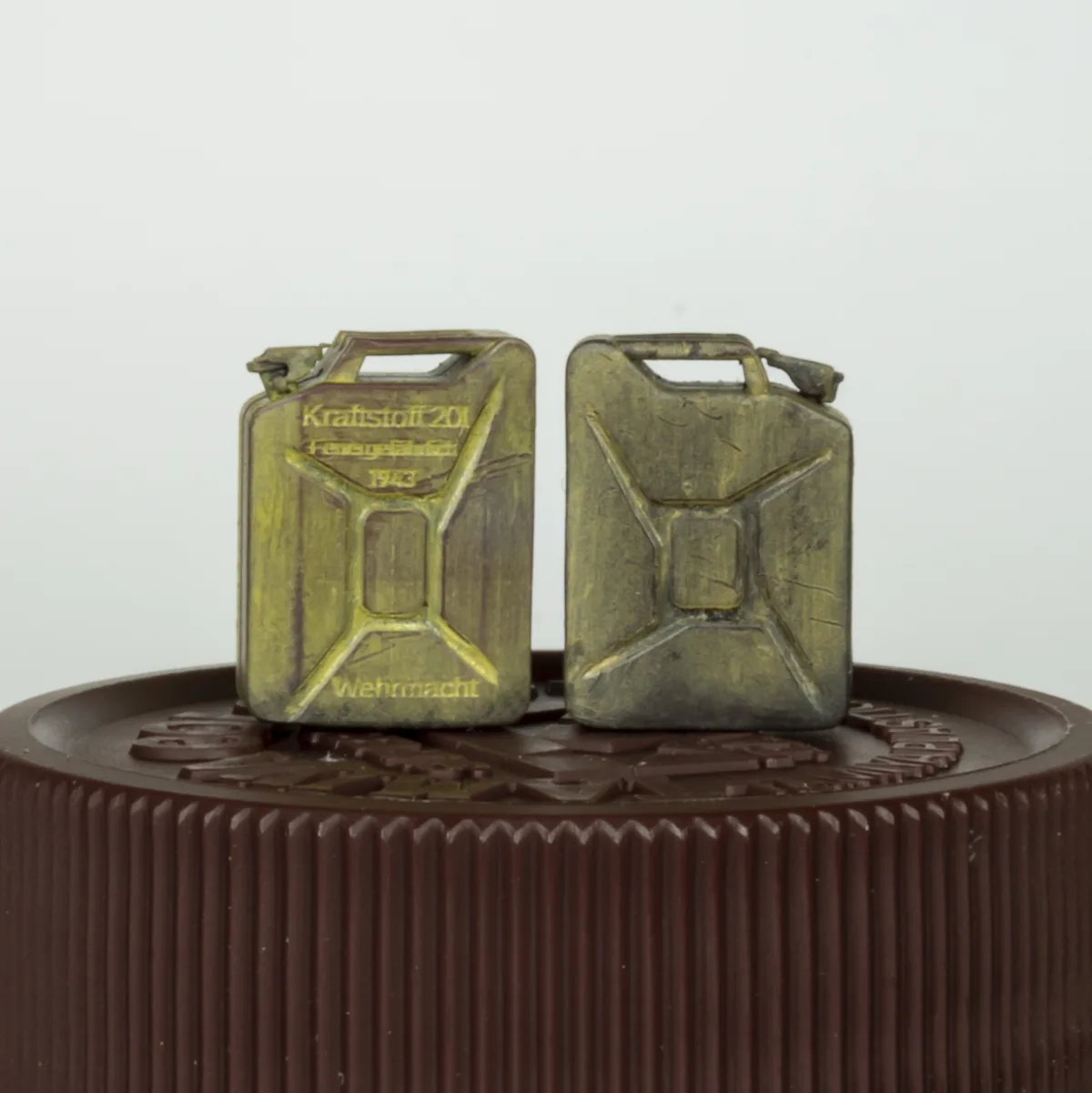 Two miniature, olive-drab metal fuel canisters sit atop a large, dark brown chocolate bar with embossed text. The canister…